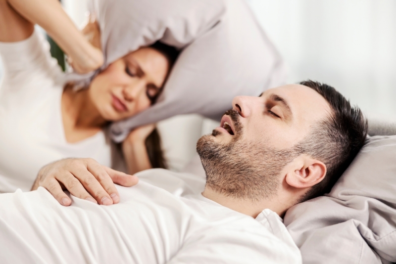 Man in bed snoring due to sleep apnea and woman covering her ears with a pillow in Calgary